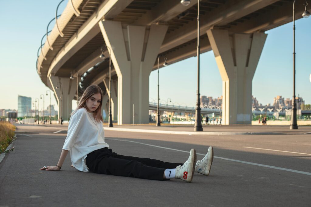 Young woman in casual streetwear sitting under an urban overpass in daylight.
